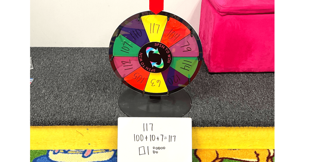 a wheel with numbers to 120 written on the spots and a whiteboard below it with 117 written in standard form, expanded form, and picture form